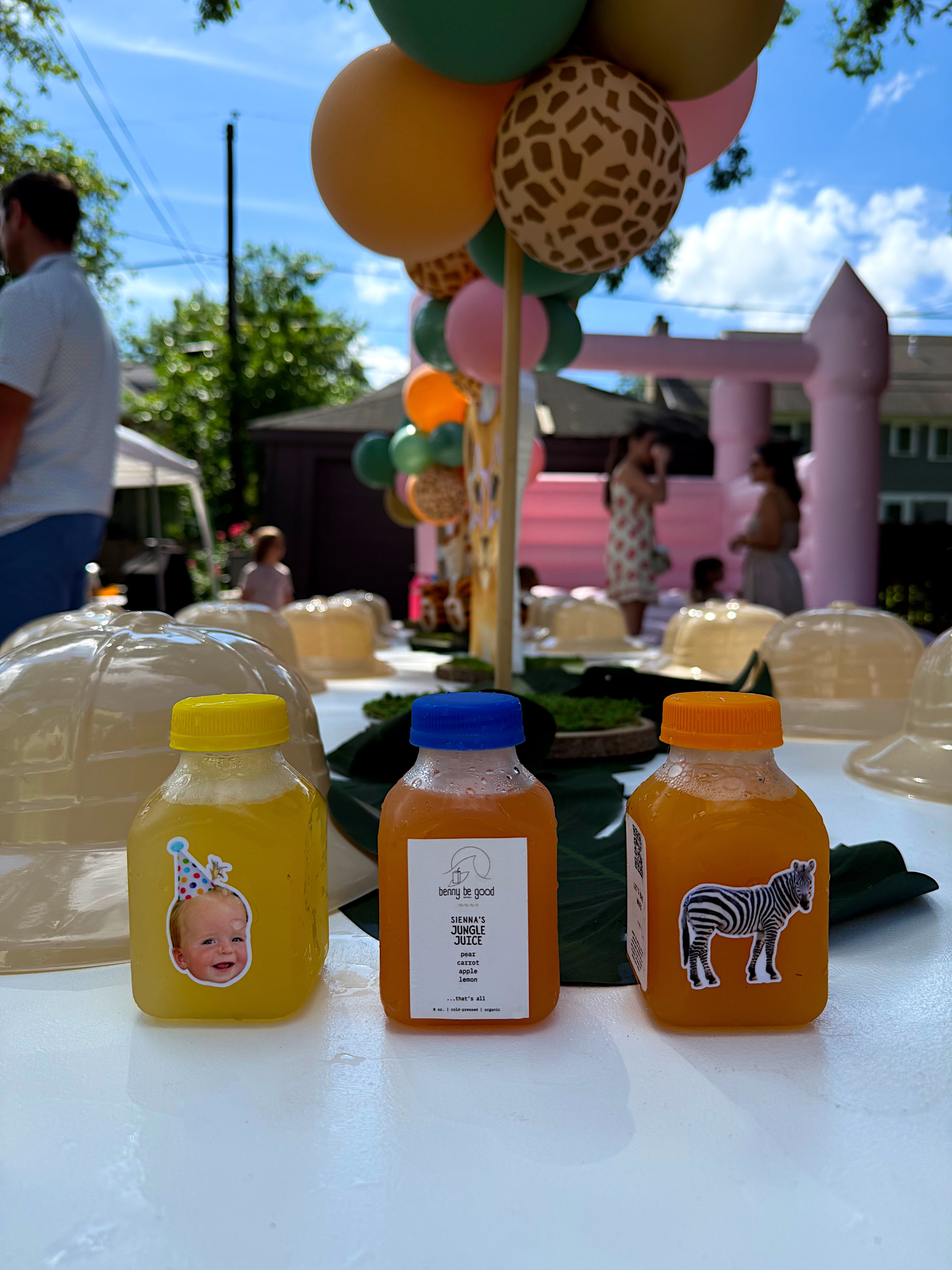 Three small bottles with colorful labels on a table with balloons and a pink inflatable structure in the background.